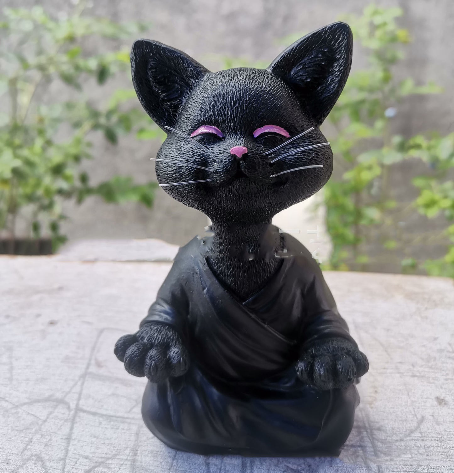 Black cat figurine with pink accents on a textured surface with greenery in the background