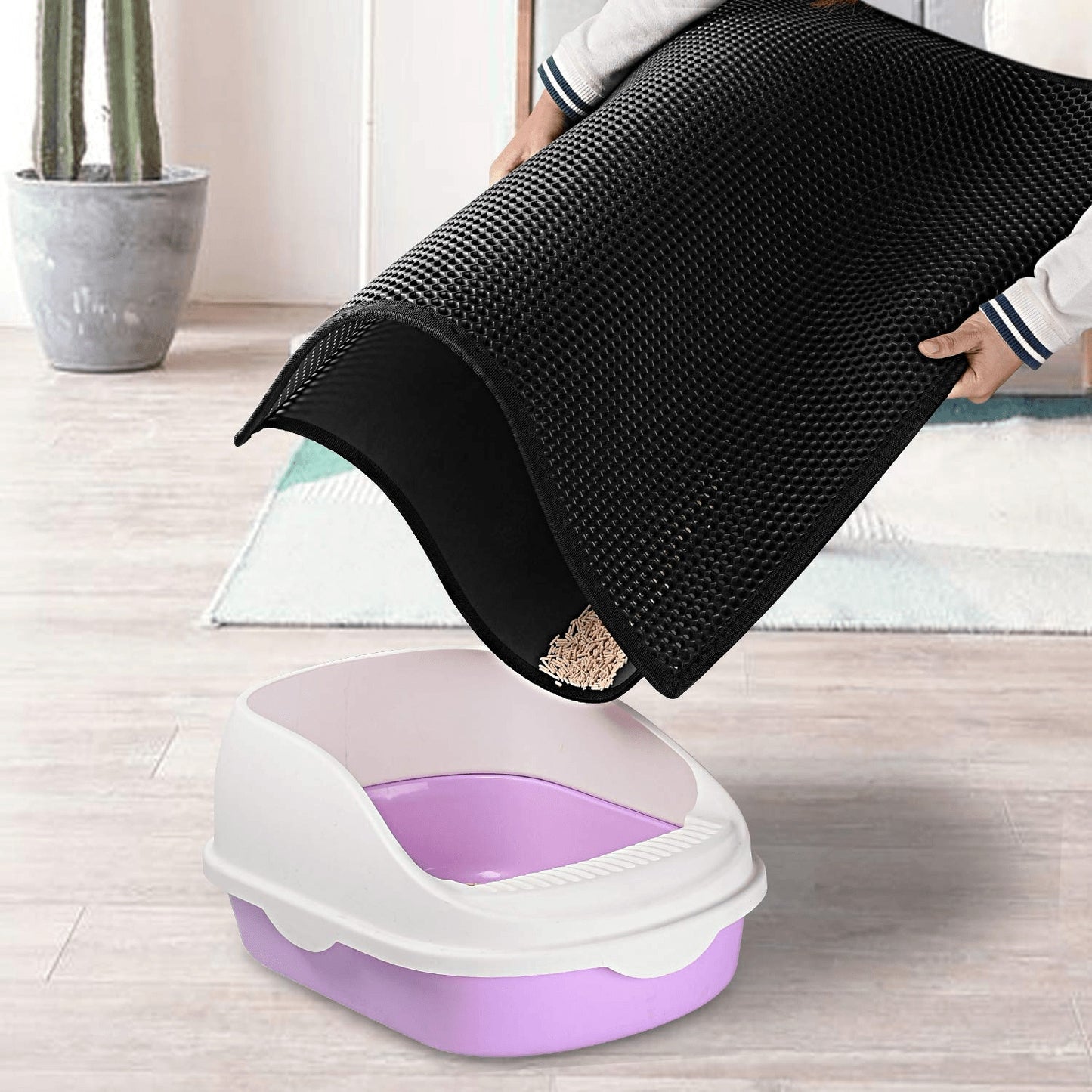 Person using a black mat to clean a purple and white litter box on a wooden floor.