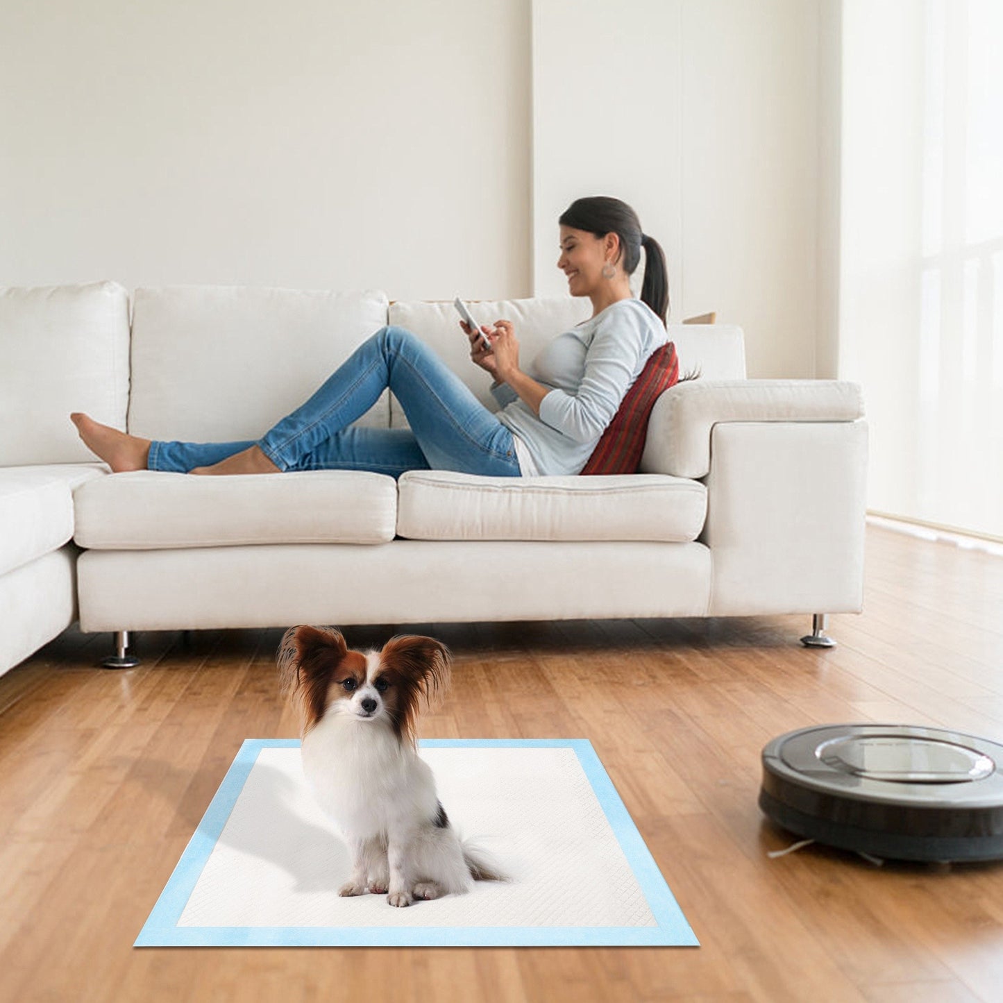 A smiling woman sitting on a couch and looking at her phone, with her dog resting calmly on a training pad beside her.