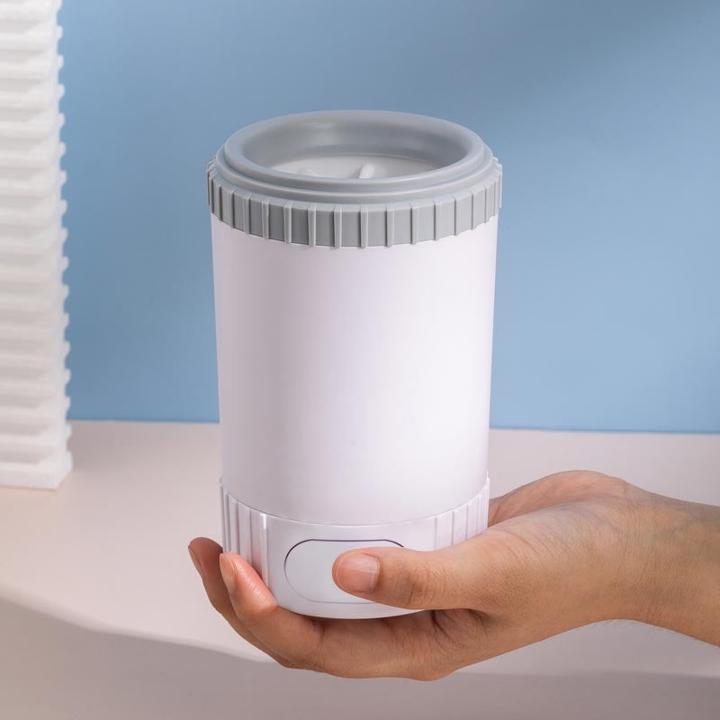 Hand holding a cylindrical white and gray dog paw cleaning device against a light blue background