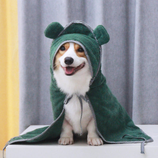 Dog wearing a green hooded towel with a bear design, sitting on a white surface against a gray curtain background.