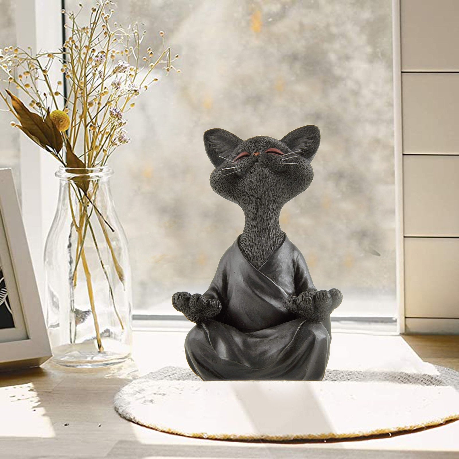Gray cat figurine in a robe meditating on a table with a vase of dried flowers in the background.