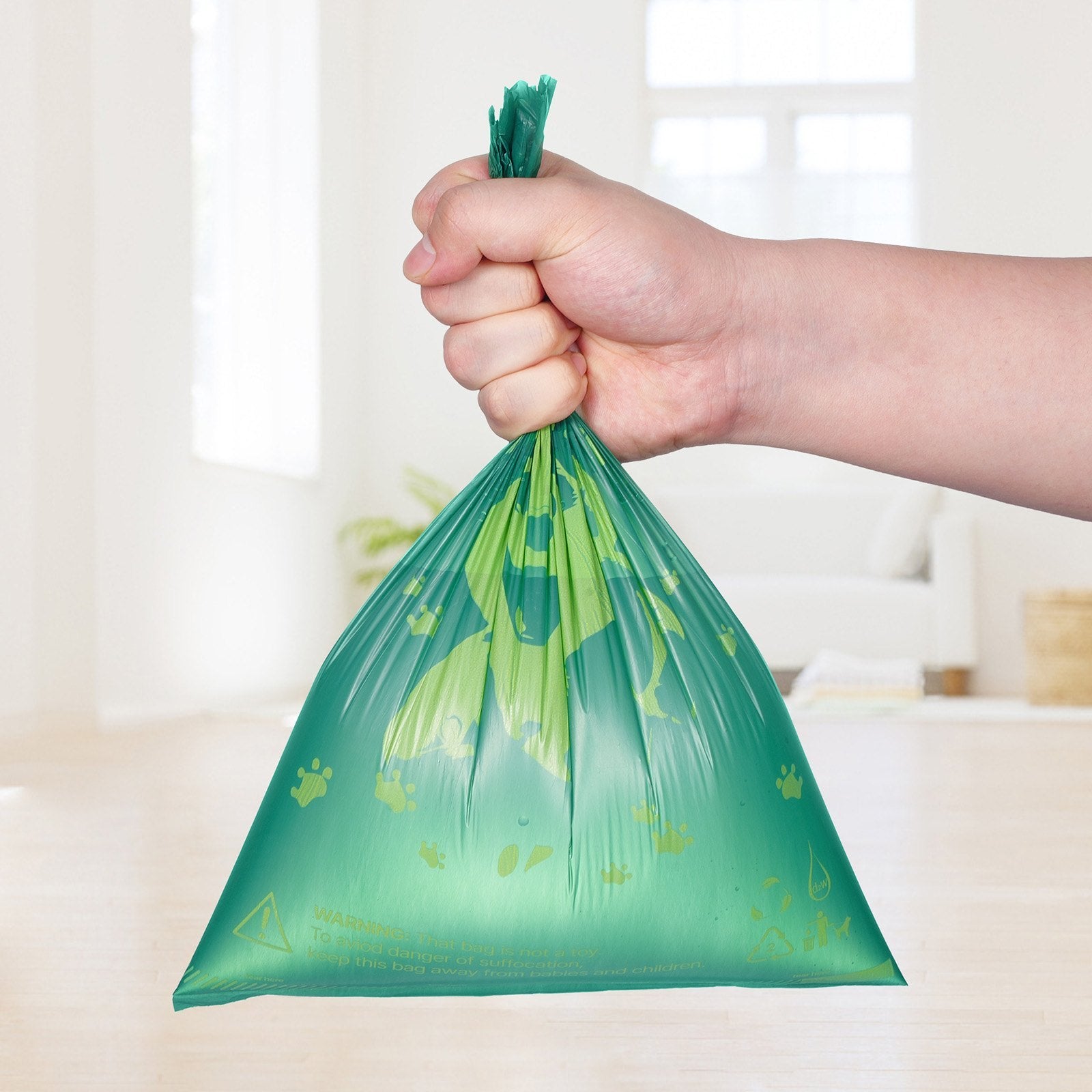 Hand holding a green plastic bag with paw prints on a blurred indoor background